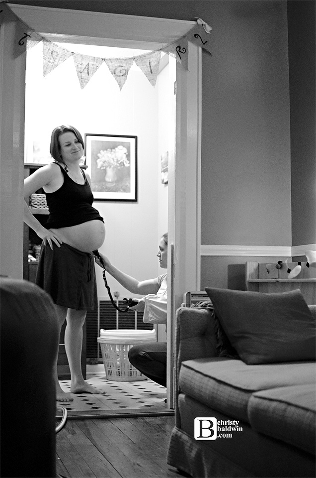 pregnant woman with belly showing with kneeling midwife listening to baby's heart tones with handheld doppler in a doorway at a home birth.