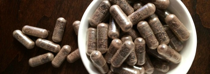 Photo of white bowl of brown pills with pills next to the bowl with brown background. Placenta pills are the main cost of placenta encapsulation. 