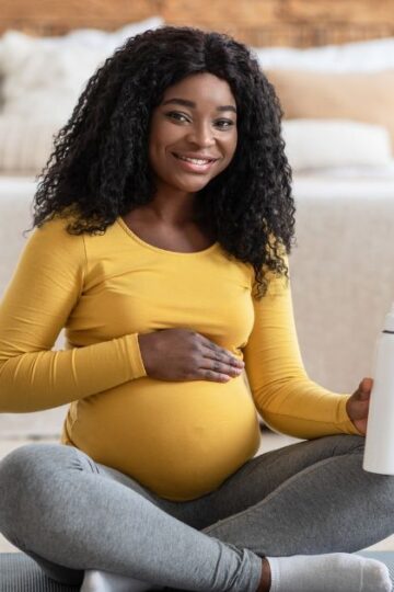 black pregnant woman with yellow shirt sitting on a yoga mat with a water bottle